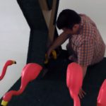 A man kneels by an art piece surrounded by four bright pink flamingoes.