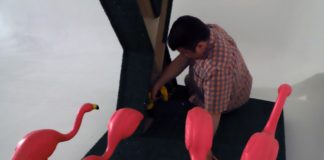 A man kneels by an art piece surrounded by four bright pink flamingoes.