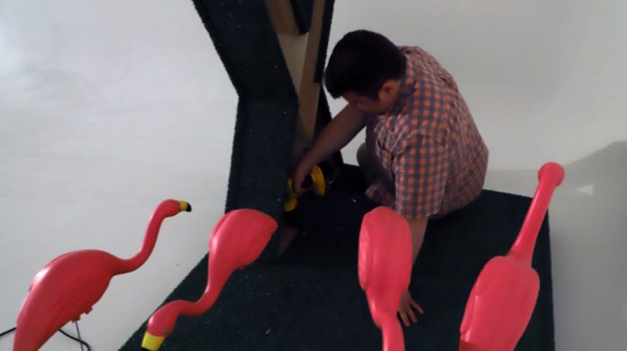 A man kneels by an art piece surrounded by four bright pink flamingoes.