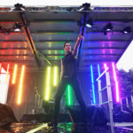 Brie Treviranus striking a pose on stage with the stage lights set to the pride rainbow.